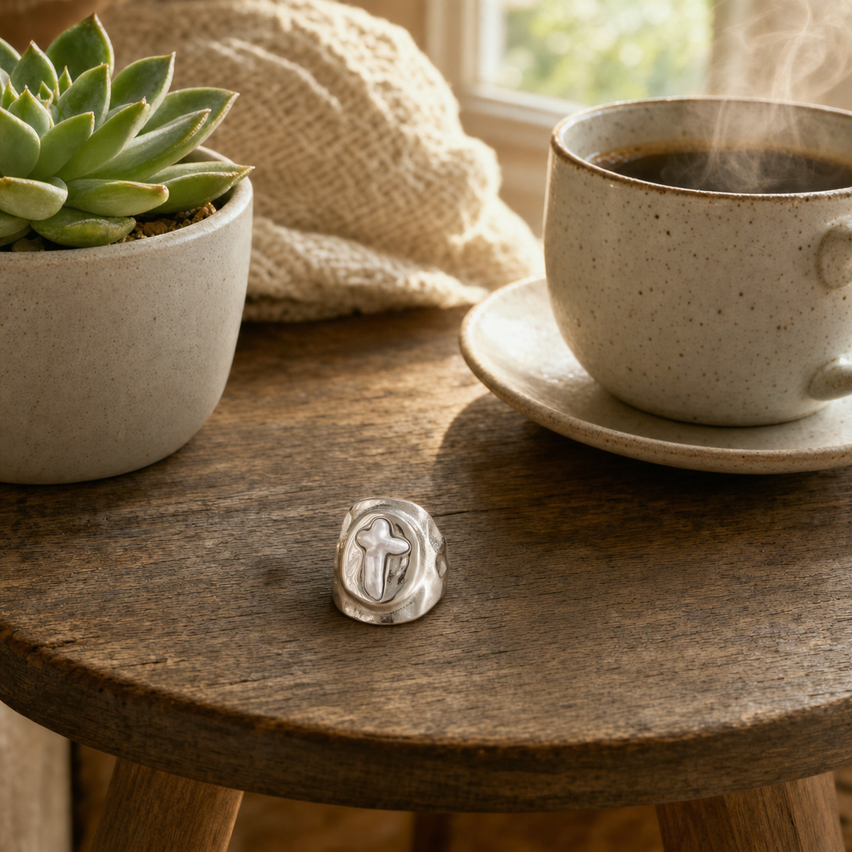 Silver tone stainless steel wide molten dome signet ring with mother of pearl cross inlay in oval recess, hypoallergenic tarnish resistant faith-inspired statement ring, displayed on a round wood side table with a succulent and coffee by The Intertwined Collective