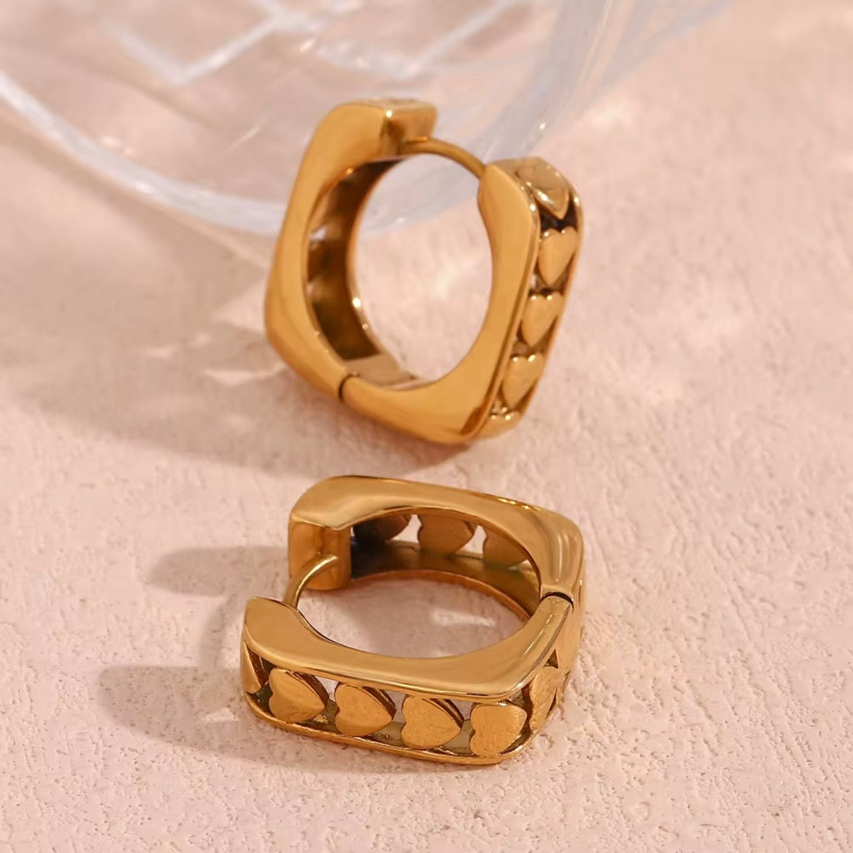 18k gold square huggie hoop earrings with embossed heart row on outer band displayed on neutral surface — hypoallergenic water-resistant tarnish-resistant stackable huggies by The Intertwined Collective, Dallas Texas