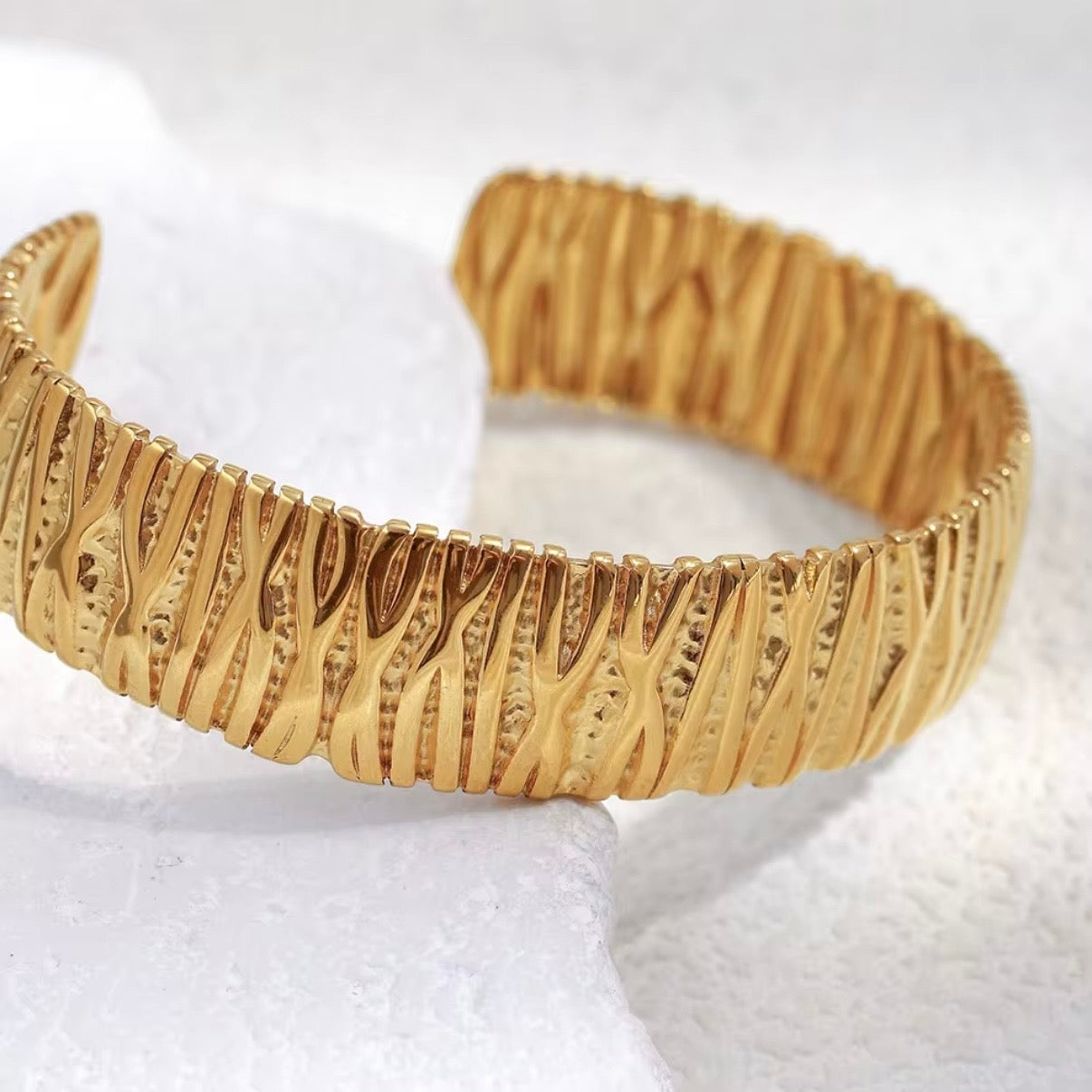 Wide gold open cuff bangle with hammered ripple texture displayed on white stone and soft neutral background – hypoallergenic water and tarnish resistant statement bracelet"