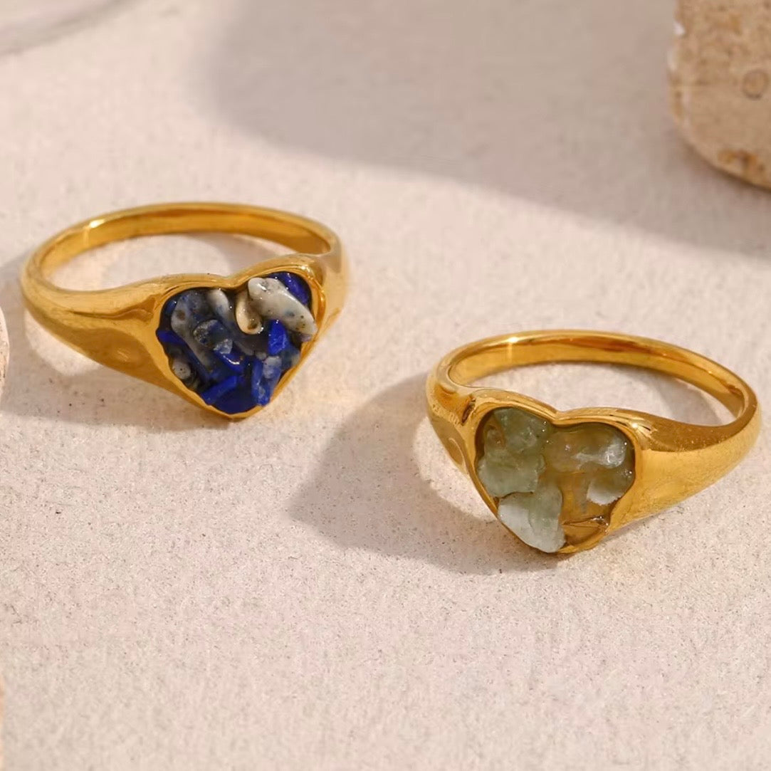 Four gold heart signet rings with crushed gemstone inlays in blue and pale green arranged on a sandy beige surface beside a seashell.