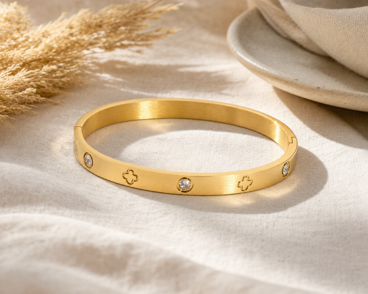 Gold bangle with clover cutout engravings and round crystal accents styled on linen with dried pampas grass