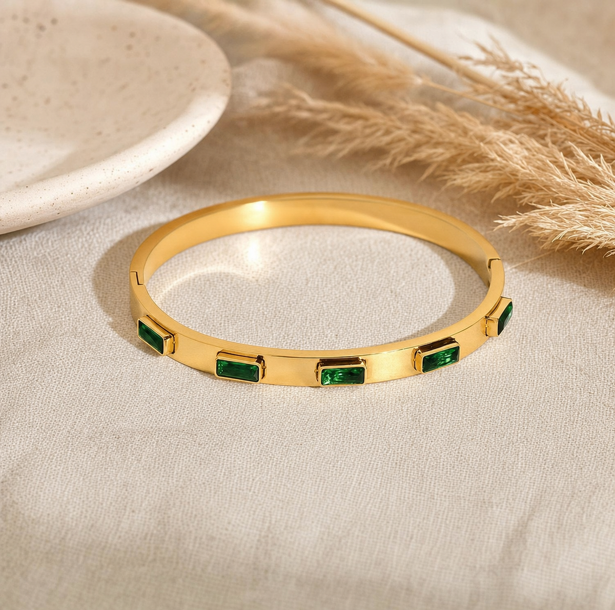 Gold bangle with emerald rectangle stones displayed on linen with dried pampas grass