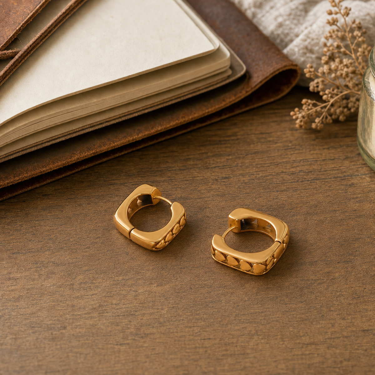 18k gold square huggie hoop earrings with embossed heart row on outer band displayed on a rustic wood desk with a leather journal and dried florals — hypoallergenic water-resistant tarnish-resistant stackable huggies by The Intertwined Collective, Dallas Texas