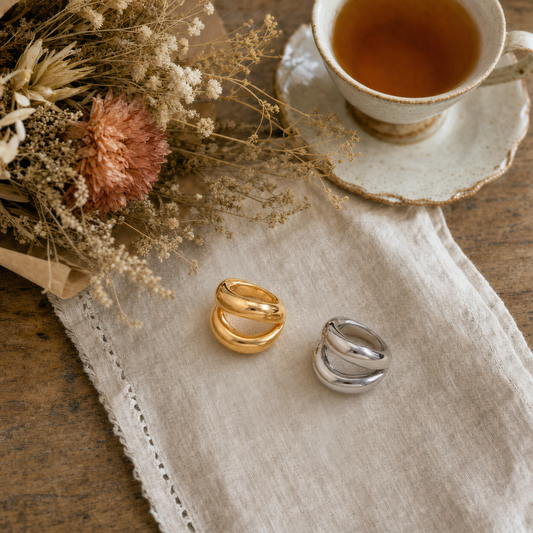 18k gold plated and silver tone stainless steel double wave dome statement ring with organic wavy sculptural silhouette and high-polish mirror finish, hypoallergenic tarnish resistant ring by, displayed on a linen napkin with dried florals and tea. The Intertwined Collective