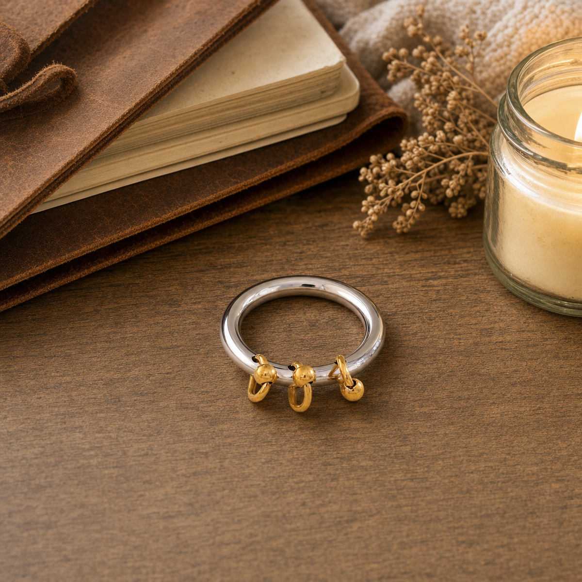 Two-tone stainless steel charm band ring with smooth silver tone band and three dangling 18k gold plated ball-and-hoop charms, hypoallergenic tarnish resistant playful statement ring by The Intertwined Collective, displayed on a rustic wood desk with a leather journal, candle, and dried florals