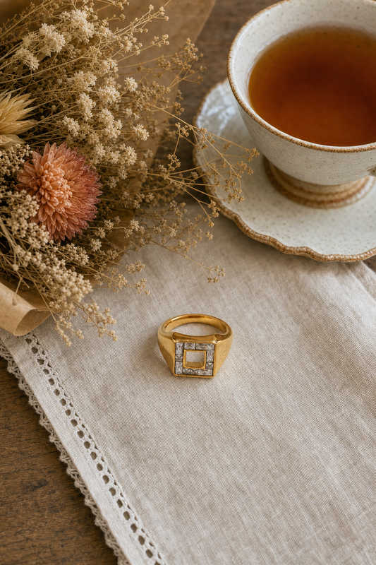 18k gold plated stainless steel wide signet ring with diamond-shaped face, baguette CZ channel-set frame and open square center cutout, hypoallergenic tarnish resistant architectural statement ring, displayed on a linen napkin with dried florals and tea by The Intertwined Collective