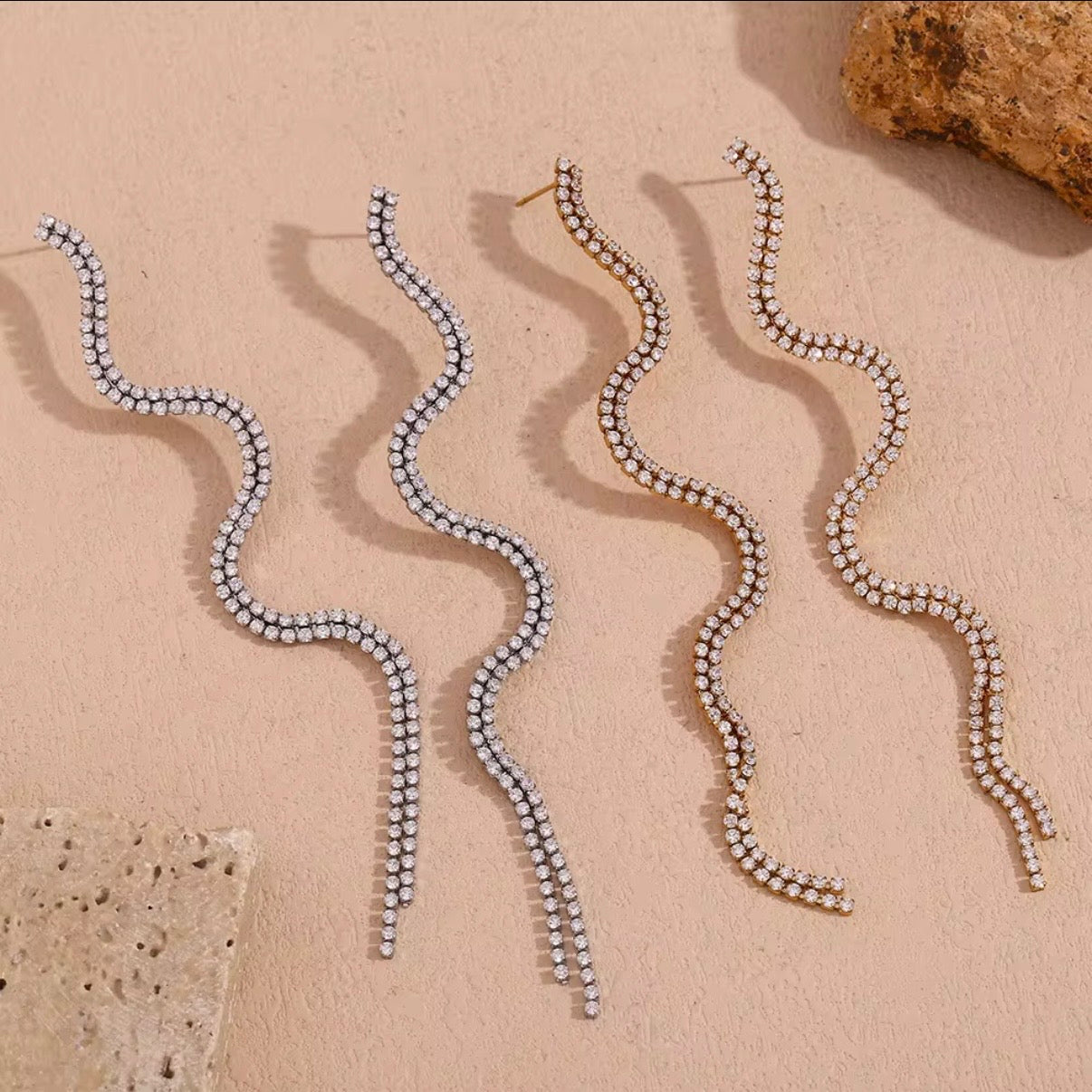 Gold and silver double-row pavé CZ crystal wave fringe drop earrings displayed on neutral stone surface — hypoallergenic water-resistant tarnish-resistant long crystal statement earrings by The Intertwined Collective, Dallas Texas