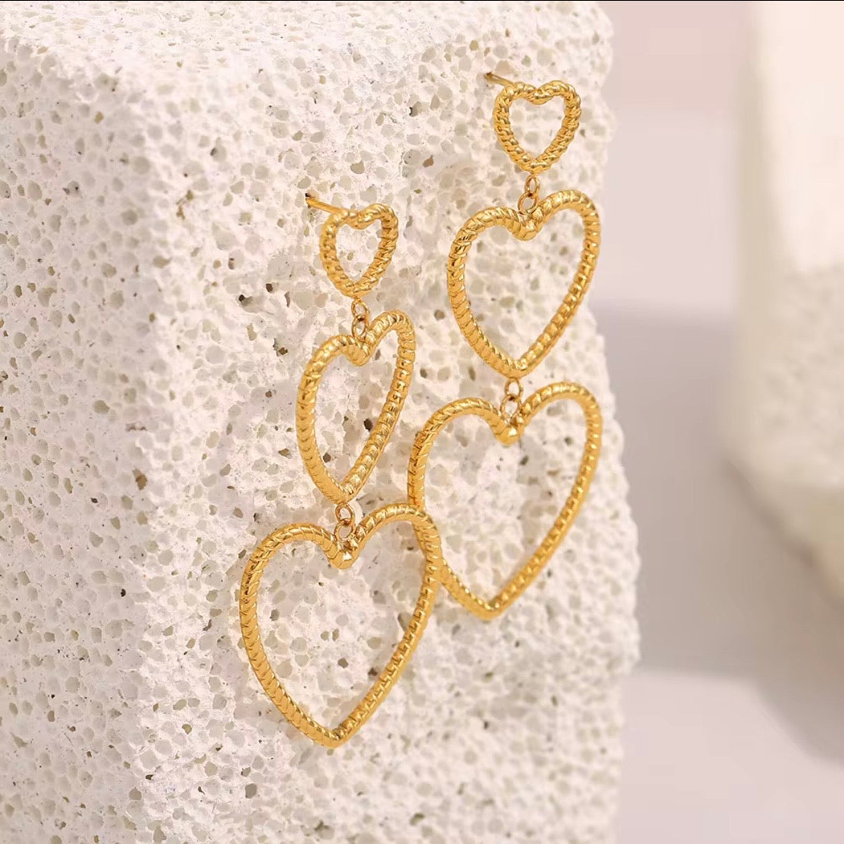 Gold triple heart drop earrings with textured rope-style open hearts displayed on white stone – lightweight hypoallergenic water- and tarnish‑resistant statement earrings