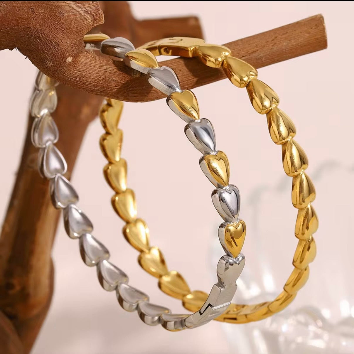 Gold and silver metal heart-link bangles made from continuous puffed hearts, stacked together on stone and wood props – hypoallergenic, water- and tarnish‑resistant heart bangles