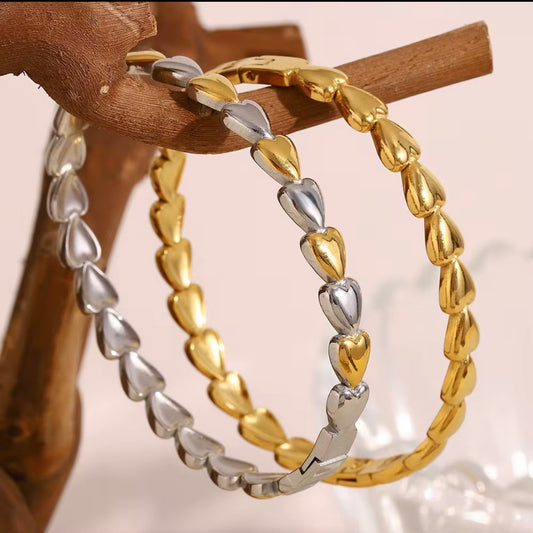 Gold and silver metal heart-link bangles made from continuous puffed hearts, stacked together on stone and wood props – hypoallergenic, water- and tarnish‑resistant heart bangles