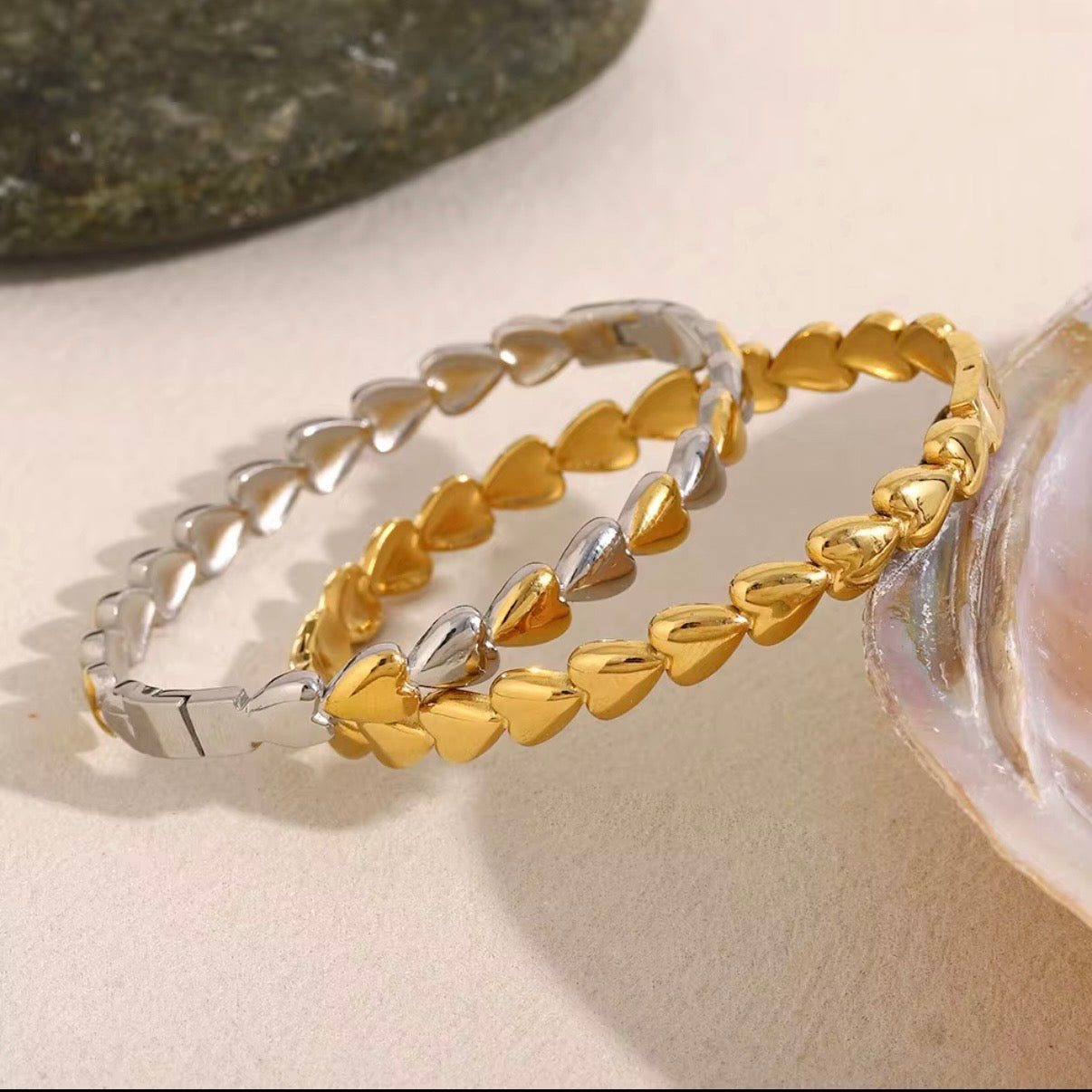 Gold and silver metal heart-link bangles made from continuous puffed hearts, stacked together on stone and wood props – hypoallergenic, water- and tarnish‑resistant heart bangles