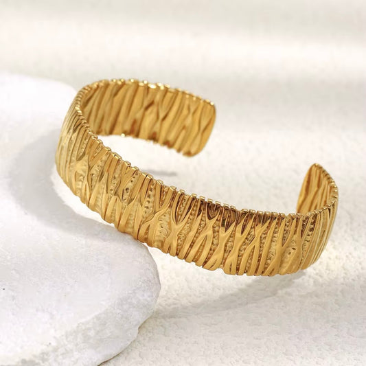 Wide gold open cuff bangle with hammered ripple texture displayed on white stone and soft neutral background – hypoallergenic water and tarnish resistant statement bracelet"