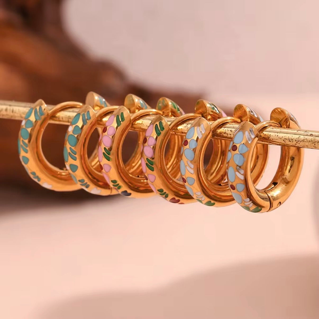 Three pairs of small gold huggie hoop earrings with colorful floral enamel in mint green, soft pink, and sky blue displayed on bar and stone – hypoallergenic water and tarnish resistant huggies