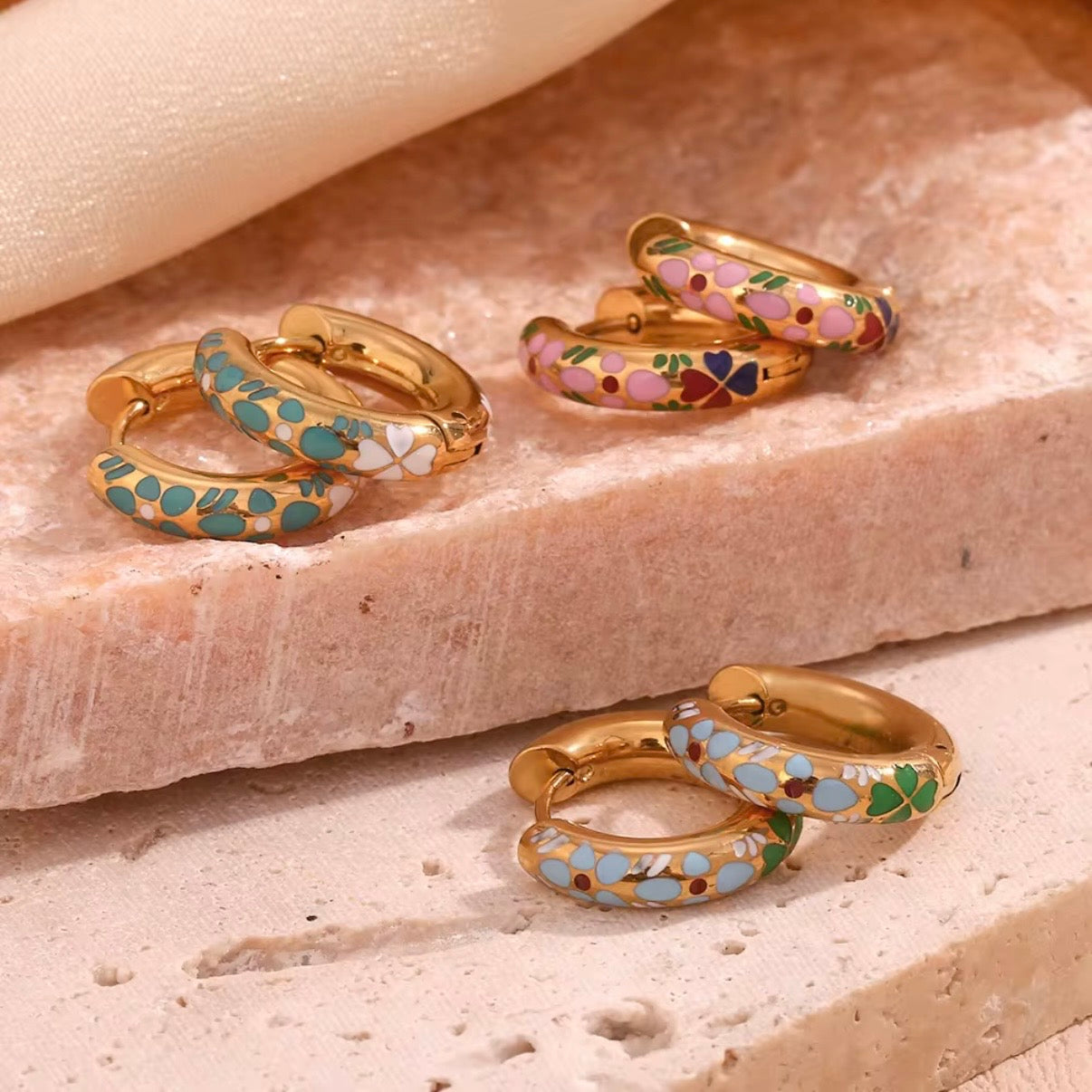 Three pairs of small gold huggie hoop earrings with colorful floral enamel in mint green, soft pink, and sky blue displayed on bar and stone – hypoallergenic water and tarnish resistant huggies
