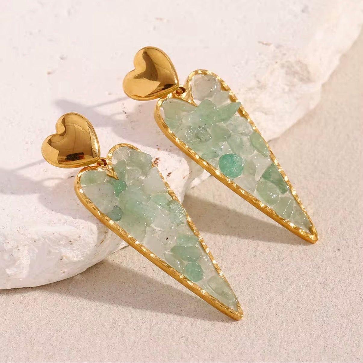 Gold heart stud earrings with elongated heart drops filled with raw green crystal chips displayed on white stone – hypoallergenic water and tarnish resistant statement earrings