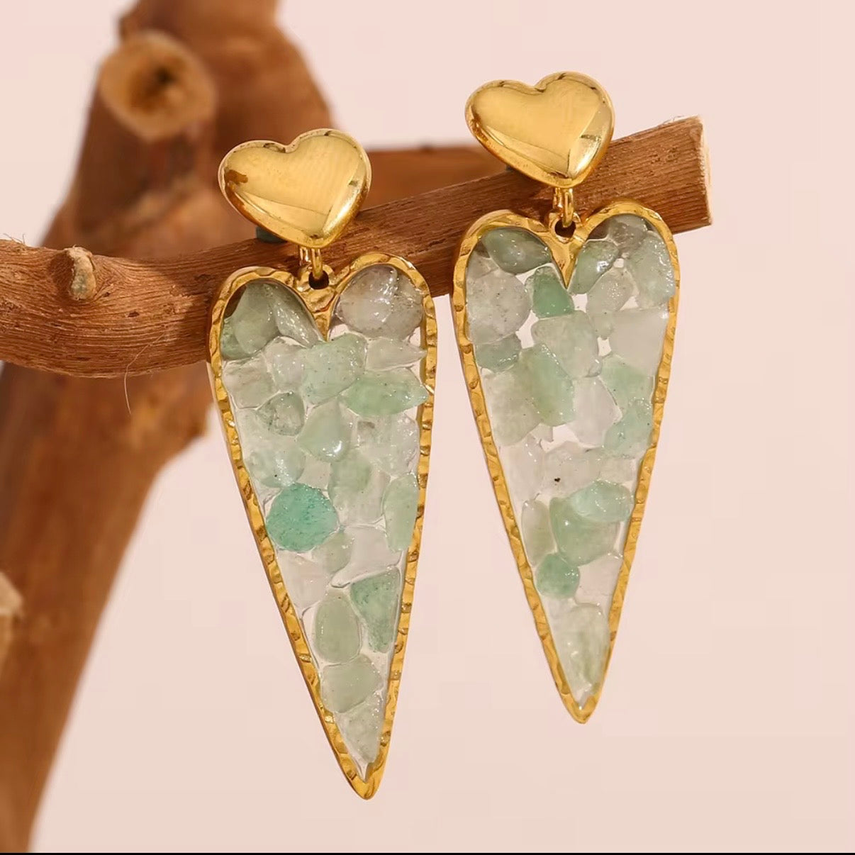 Gold heart stud earrings with elongated heart drops filled with raw green crystal chips displayed on white stone – hypoallergenic water and tarnish resistant statement earrings