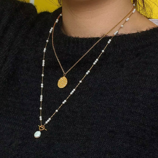 Layered gold necklaces on model wearing black sweater, featuring pearl station chain with drop pearl pendant and shorter gold oval medallion – hypoallergenic water and tarnish resistant layered necklace set