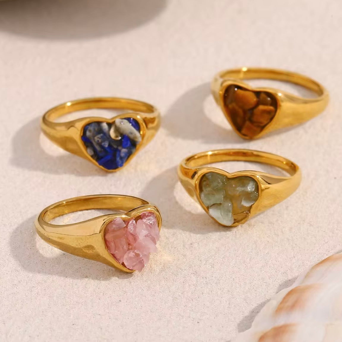 Gold heart signet rings with raw natural stone inlays in lapis lazuli, tiger's eye, rose quartz, and jade displayed on sandy surface – hypoallergenic crystal statement rings