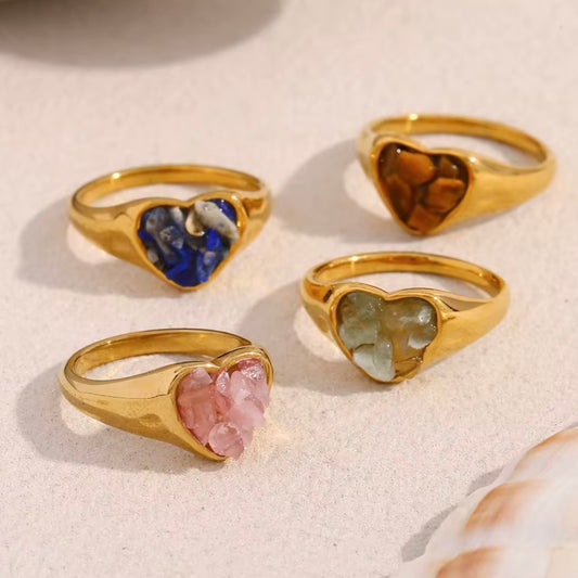 Gold heart signet rings with raw natural stone inlays in lapis lazuli, tiger's eye, rose quartz, and jade displayed on sandy surface – hypoallergenic crystal statement rings