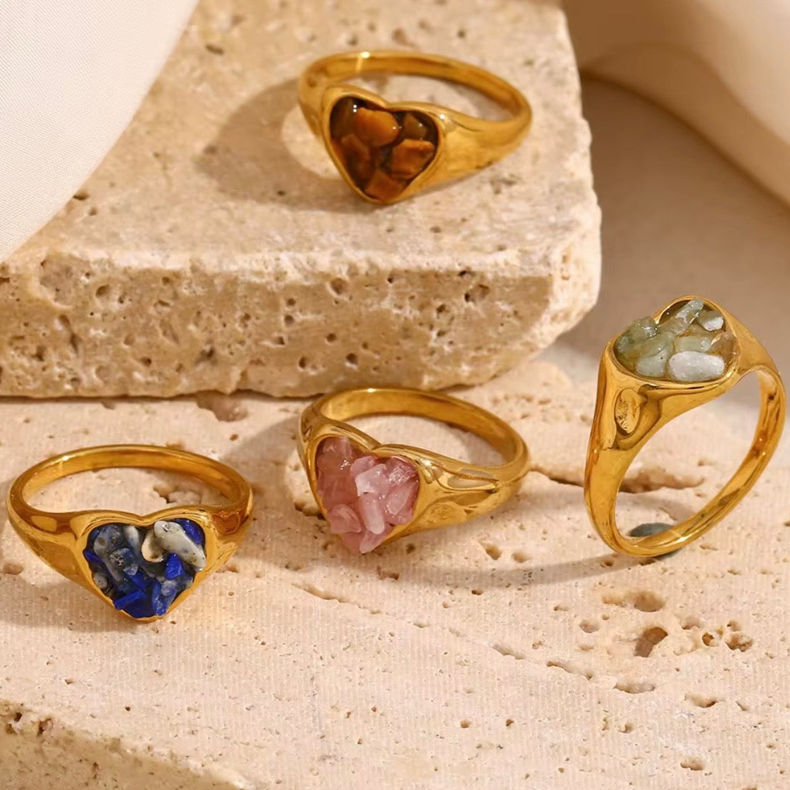 Gold heart signet rings with raw natural stone inlays in lapis lazuli, tiger's eye, rose quartz, and jade displayed on sandy surface – hypoallergenic crystal statement rings