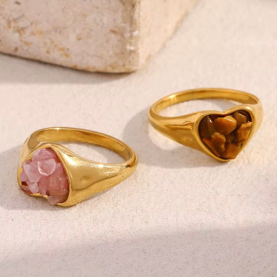 Four gold heart signet rings with crushed gemstone inlays in  amber and soft pink, arranged on a sandy beige surface beside a seashell.