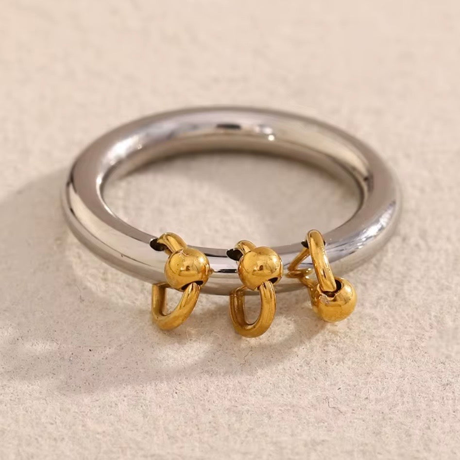 Two-tone stainless steel charm band ring with smooth silver tone band and three dangling 18k gold plated ball-and-hoop charms, hypoallergenic tarnish resistant playful statement ring by The Intertwined Collective
